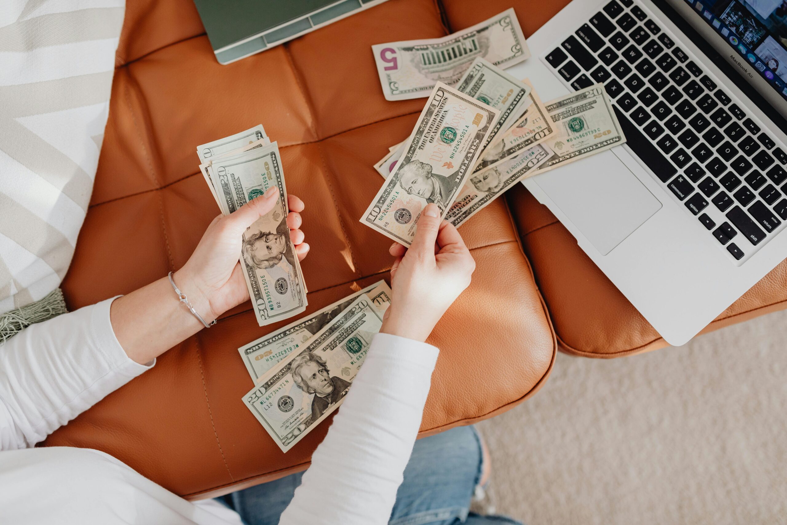 A person counts US dollar bills on a couch with a laptop, symbolizing financial planning.Earn Money Online